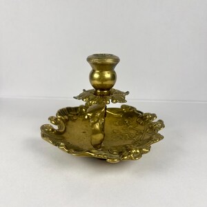 May include: A vintage brass candlestick holder with a decorative, ornate design. The candlestick features a round base with a floral pattern and a central cup for holding a candle. The holder is made of brass and has a warm, golden color.