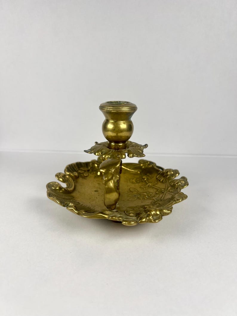 May include: A vintage brass candlestick holder with a decorative, ornate design. The candlestick features a round base with a floral pattern and a central cup for holding a candle. The holder is made of brass and has a warm, golden color.
