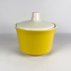 Op de afbeelding: Een gele keramische suikerpot met een crèmekleurig deksel en een klein handvat. De kom heeft een afgeronde vorm en een gladde, glanzende afwerking. Het deksel past goed op de bovenkant en zorgt voor een goede afdichting. Het is een keukengerei.