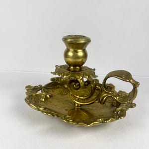 May include: Ornate brass candlestick holder with a decorative handle and floral accents. The candle holder has a round base and a detailed design, perfect for adding a touch of vintage charm to any space.