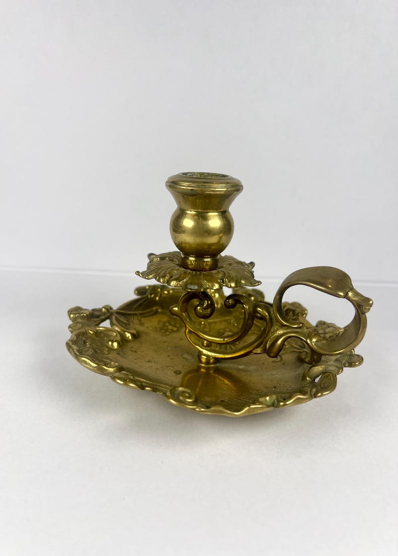 May include: Ornate brass candlestick holder with a decorative handle and floral accents. The candle holder has a round base and a detailed design, perfect for adding a touch of vintage charm to any space.