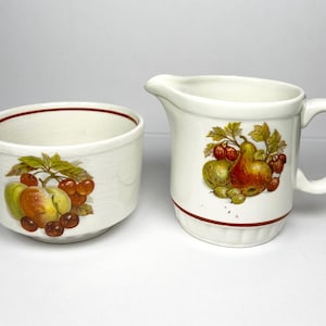 May include: A cream-coloured ceramic creamer jug and sugar bowl set. The jug has a handle and spout, and the bowl is round. Both pieces feature a fruit design with pears, cherries, and apples, and a brown trim.