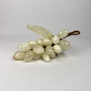Peut inclure: Un ensemble décoratif de raisins en pierres translucides blanc cassé. Les raisins ovales sont attachés à une tige marron avec une seule feuille. L'arrangement est posé sur un fond blanc.