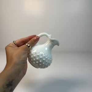 May include: A small white milk glass creamer featuring a hobnail texture across the rounded body. The vessel has a curved handle and a small pouring spout. The surface of the glass is opaque and glossy, reflecting light across the raised bumps.