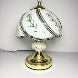 Peut inclure: Une lampe de table vintage avec une base en laiton et un abat-jour blanc à motifs floraux. La lampe comporte un fleuron décoratif et une section centrale en céramique avec des accents floraux roses. L'abat-jour mesure environ 25 cm de diamètre.