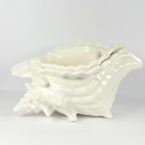 Puede incluir: Un cuenco decorativo de cerámica en forma de concha de color crema. El cuenco tiene una superficie texturizada con un borde acanalado y se apoya sobre tres pequeños pies. El diseño imita la forma natural de una concha.