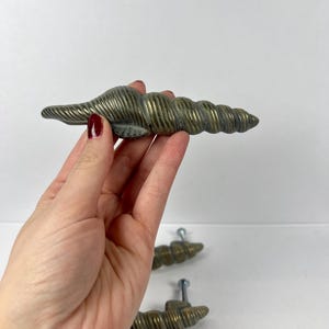 Op de afbeelding: Een set messingkleurige schelpvormige kastknoppen. De knoppen hebben een getextureerd, geribbeld oppervlak en zijn gemonteerd met zilveren schroeven. De afbeelding toont één knop die wordt vastgehouden en drie andere op de achtergrond.