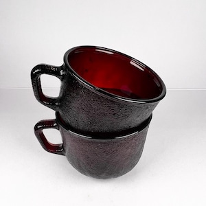 Könnte beinhalten: Zwei gestapelte, strukturierte, dunkelrote Glasbecher mit Henkeln. Das Innere der Becher ist leuchtend rot und kontrastiert mit dem dunkleren Äußeren. Die Becher sind wahrscheinlich für Getränke.