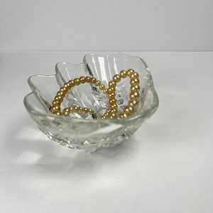 May include: A clear, textured glass bowl in a flower shape holds a strand of golden beads. The bowl has a scalloped edge and sits on a white surface. The beads are a warm, light gold colour.