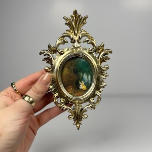 May include: An oval picture frame in a gold colour, with an ornate, scrolling design. The frame holds a small painting with green and brown tones. The frame is being held by a person.