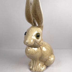 Op de afbeelding: Een keramieken konijnfiguur met lange oren en grote, groene ogen. Het konijn is beige met bruine spikkels en een glanzende afwerking. Het beeldje is een decoratief item, geschikt voor woondecoratie.