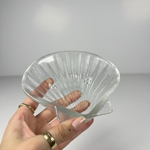 May include: A clear glass dish shaped like a scallop shell with a textured, ribbed surface. The dish features three small, raised circular feet on the interior base, providing stability. The glass is translucent and reflects light along its ridges.