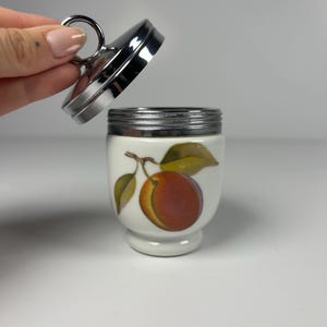 May include: A white porcelain egg coddler with a chrome metal lid featuring a ring handle. The body of the coddler is decorated with a colourful illustration of a peach with green leaves on a stem. The lid is shown detached from the base.