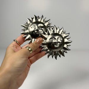 Peut inclure: Deux ornements solaires en métal argenté de forme circulaire, présentant des visages en relief avec des yeux fermés et des bouches souriantes, entourés de rayons pointus. Les soleils ont une finition lisse, réfléchissante et un design céleste.