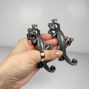 May include: Two silver-toned metal cabinet pulls shaped like lizards. Each lizard figure features a textured, weathered finish with a curled tail and splayed feet, designed to function as a handle for furniture or cabinetry hardware.