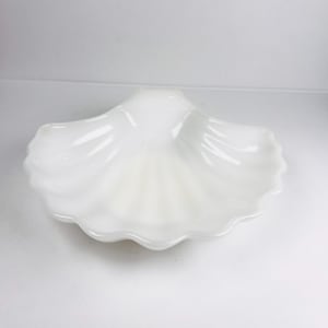 May include: A white, opalescent seashell-shaped dish. The dish has a scalloped edge and a smooth, glossy surface. The interior of the dish has a ribbed texture, and the overall design is reminiscent of a natural seashell.