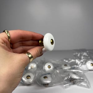 Peut inclure: Boutons de meuble blancs et dorés. Les boutons ont une face ronde en céramique blanche avec un centre et une base dorés. Plusieurs boutons sont visibles dans un sac en plastique transparent.