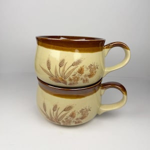 Peut inclure: Deux tasses en céramique beiges empilées avec des bords et des anses marron. Chaque tasse présente un motif de blé et de fleurs dans des tons de marron. Les tasses sont rondes avec une large ouverture.