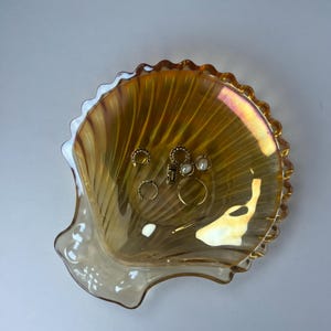 Peut inclure: Un plat décoratif en forme de coquillage en verre irisé avec un bord festonné. Le plat contient plusieurs bagues et boucles d'oreilles dorées. Le verre est de couleur ambre chaud avec des reflets roses et jaunes.