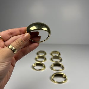 May include: A set of gold-toned metal napkin rings with a smooth, rounded, dome-shaped top surface. The collection includes one ring held up for display and five additional matching rings arranged in two rows on a plain surface.
