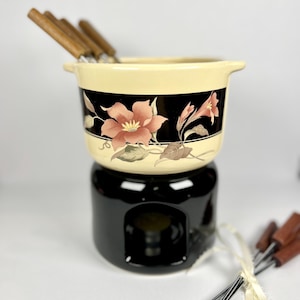 Peut inclure: Un service à fondue vintage avec un pot en céramique crème orné d'un motif floral rose et noir. L'ensemble comprend une base noire et des fourchettes à fondue avec des manches en bois. Un ajout charmant.