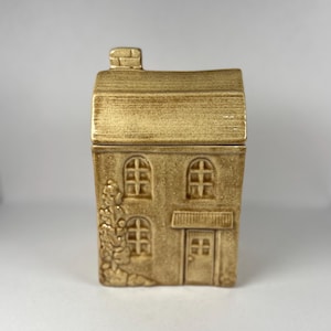 Peut inclure: Un récipient en céramique beige en forme de maison avec un couvercle amovible. La maison présente des fenêtres cintrées, une porte d'entrée et une cheminée. L'extérieur a une finition texturée et un motif floral sur le côté.