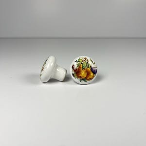 Puede incluir: Dos pomos de armario de cerámica blanca. Cada pomo presenta un diseño pintado a mano de varias frutas, incluyendo peras y ciruelas, en tonos amarillos, morados y verdes. Los pomos se colocan sobre un fondo blanco liso, resaltando su naturaleza decorativa.