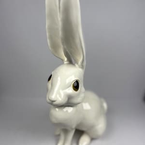 Könnte beinhalten: Eine weiße Keramik-Hasenfigur mit langen, aufrechten Ohren und braunen Augen. Der Hase sitzt, die Vorderpfoten auf dem Boden. Die Figur hat eine glänzende Oberfläche und ist ein dekorativer Artikel.
