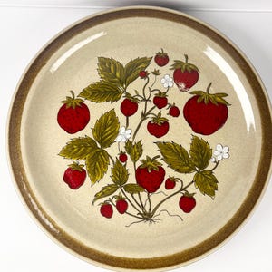 Peut inclure: Assiette ronde en céramique avec un fond beige et un bord marron. L'assiette présente un motif de fraisier avec des fraises rouges, des feuilles vertes et de petites fleurs blanches. Les fraises sont de tailles variables, et les feuilles ont un aspect détaillé et naturaliste.