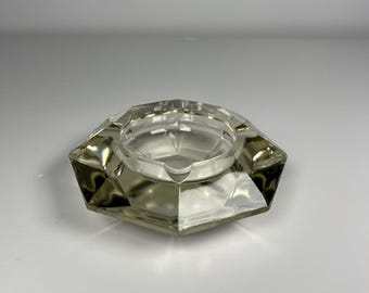 May include: A clear, faceted glass ashtray with a hexagonal outer shape and a circular central basin. The thick glass features geometric, angular cuts along the exterior edges, creating a reflective surface that catches light from multiple angles.
