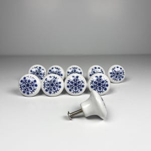 May include: A collection of white ceramic cabinet knobs with a blue floral design. The knobs are round with a decorative pattern and a metal screw for attachment. The knobs are a decorative hardware item.
