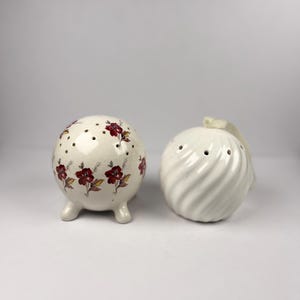 Pode incluir: Um par de saleiros e pimenteiros de cerâmica. Um é um saleiro redondo, esbranquiçado, com um padrão floral e três pés pequenos. O outro é um pimenteiro branco, com design em espiral e uma pequena pega.