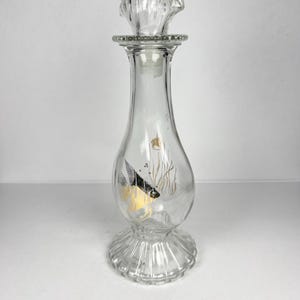 Peut inclure: Une bouteille en verre transparent avec un bouchon décoratif. La bouteille présente un motif de poisson avec des accents noirs et jaunes. La base et le bouchon ont un design côtelé.