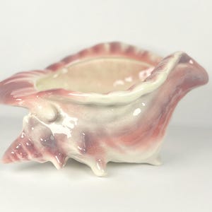 Peut inclure: Un pot en céramique en forme de coquillage dans les tons de rose, de lavande et de blanc. Le pot a une finition brillante et un extérieur texturé et détaillé, avec un intérieur lisse. Il présente une large ouverture et est soutenu par trois petits pieds.