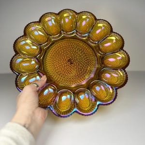 Pode incluir: Uma travessa de vidro âmbar iridescente com um design de girassol. A travessa tem uma área circular central com um padrão texturizado e é rodeada por 12 compartimentos ovais. A borda tem uma borda ondulada.
