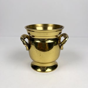 Peut inclure: Petit vase en laiton doré brillant, avec deux anses décoratives. Le vase a un corps arrondi et un bord évasé, reposant sur un petit socle. Le laiton a une finition polie.