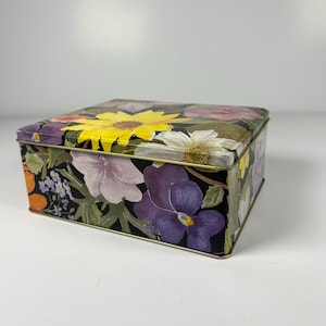 May include: A rectangular metal storage tin featuring a vibrant floral pattern on a black background. The lid and sides are decorated with painted illustrations of yellow sunflowers, purple pansies, white daisies, and orange blossoms.