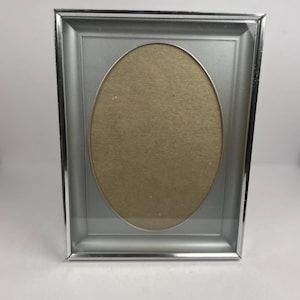 Peut inclure: Un cadre photo argenté avec une ouverture ovale. Le cadre a une forme rectangulaire avec un bord biseauté et un intérieur gris clair. Conçu pour contenir une photographie ou une œuvre d'art.