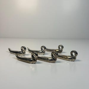 Peut inclure: Un ensemble de cinq repose-couverts en métal argenté en forme de cygne. Chaque pièce présente un corps long et fin avec un cou courbé et une tête définie, conçue avec une finition métallique lisse et réfléchissante et une silhouette élancée.