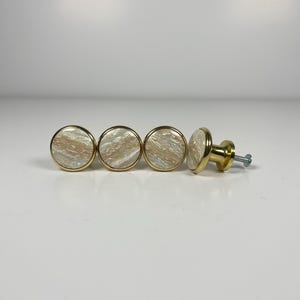 Peut inclure: Quatre boutons de placard ronds avec un bord en métal doré poli et une incrustation circulaire présentant un motif marbré tourbillonnant dans des tons crème, beige et blanc. Chaque bouton a une face lisse et une base en métal doré avec vis.
