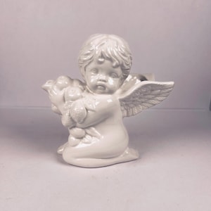 Puede incluir: Figura de querubín de cerámica blanca, arrodillado con alas y sosteniendo un racimo de fruta. El querubín tiene rasgos faciales detallados y cabello rizado. La figura es un artículo decorativo, posiblemente para la decoración del hogar o como objeto de colección.