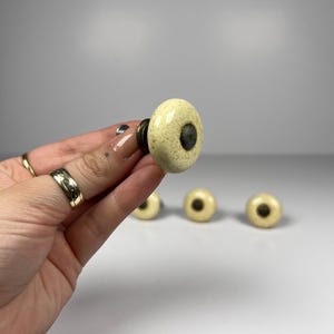 May include: A set of round ceramic cabinet knobs with a cream-coloured speckled finish and a dark metallic centre insert. Each knob features a smooth, glossy surface and a sturdy metal base, designed for attachment to furniture or cabinetry doors.