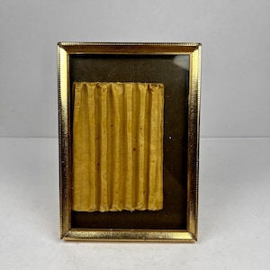 May include: A small, rectangular gold-coloured picture frame with a textured, corrugated piece of yellow paper inside. The paper is centred on a dark brown background. The frame has a simple, elegant design.