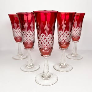 Peut inclure: Cinq flûtes à champagne rouge rubis avec des pieds et des bases transparents. La partie supérieure de chaque verre présente un motif en losanges avec des lignes verticales et horizontales. Les flûtes sont disposées en rang, mettant en valeur leur design élégant.