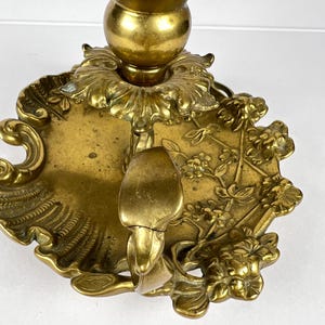 May include: A vintage brass candlestick holder with intricate floral and leaf detailing. The candle holder features a round base with a decorative edge and a central cup to hold a candle. The brass has a warm, aged patina.