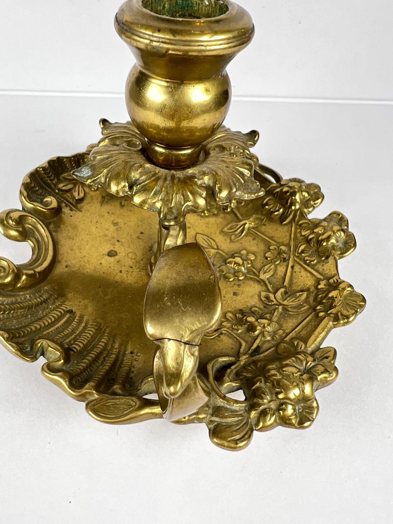 May include: A vintage brass candlestick holder with intricate floral and leaf detailing. The candle holder features a round base with a decorative edge and a central cup to hold a candle. The brass has a warm, aged patina.