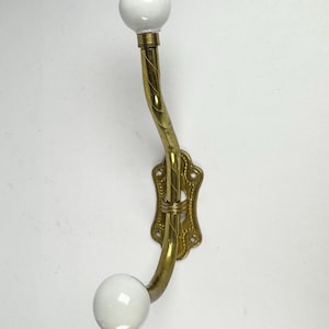Peut inclure: Patère murale vintage en laiton avec un design incurvé. Le crochet comporte deux extrémités sphériques en céramique blanche et une plaque arrière décorative en laiton. Pièce classique pour suspendre manteaux ou accessoires.