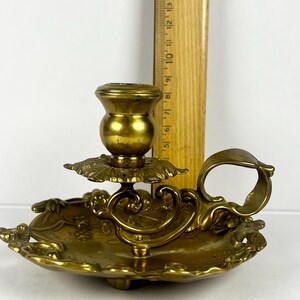 May include: Antique brass candlestick holder with a floral design. The candle holder has a round base with a handle and a decorative floral pattern. The candlestick is approximately 5 inches tall.