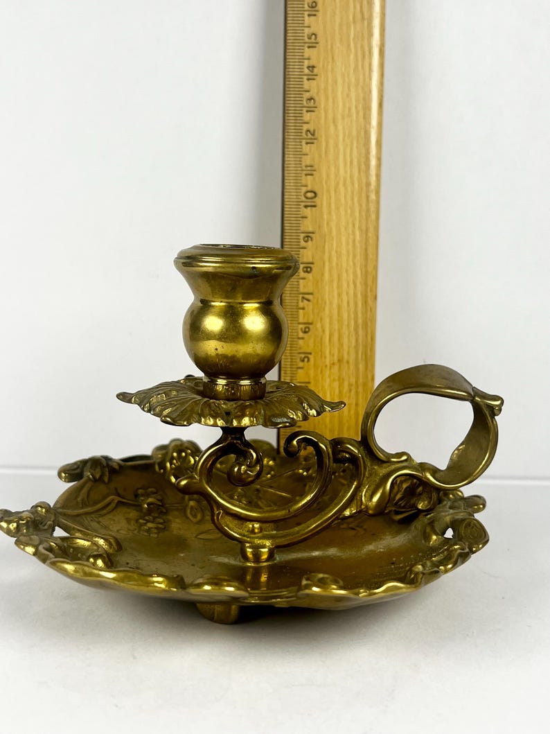 May include: Antique brass candlestick holder with a floral design. The candle holder has a round base with a handle and a decorative floral pattern. The candlestick is approximately 5 inches tall.