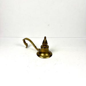 Peut inclure: Une petite lampe à huile antique en laiton. Elle possède une poignée incurvée et un sommet conique. La lampe a une base en forme de cloche et un bec verseur pour la mèche. Le laiton est de couleur dorée chaude, sur fond blanc.